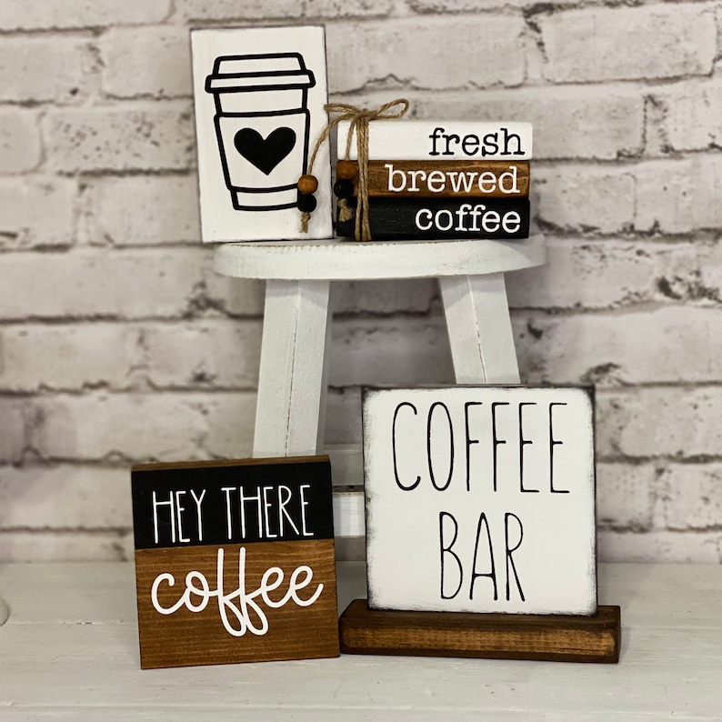 Może przedstawiać: Zestaw czterech drewnianych tabliczek z czarno-białym napisem. Tabliczki głosi: "fresh brewed coffee", "Hey there coffee" i "Coffee Bar". Tabliczki są umieszczone na białym stołku z małą drewnianą tabliczką z czarno-białym rysunkiem filiżanki kawy z sercem.