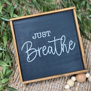 May include: A square wooden sign with a black painted surface and a natural wood frame. The text "JUST Breathe" is printed in white lettering on the center of the black surface. The sign features a clean, simple design with visible wood grain.