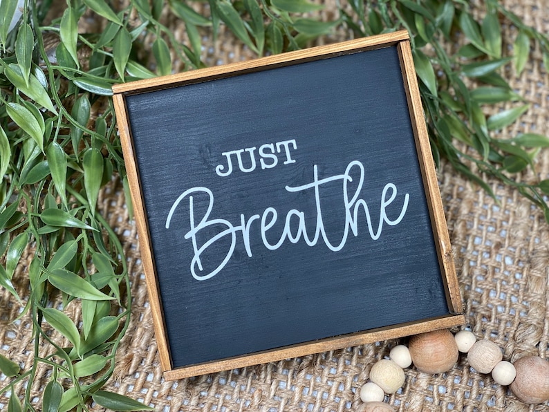 May include: A square wooden sign with a black painted surface and a natural wood frame. The text "JUST Breathe" is printed in white lettering on the center of the black surface. The sign features a clean, simple design with visible wood grain.