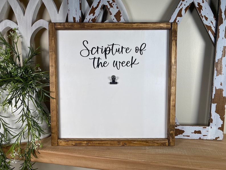 May include: A white wooden sign with a brown frame and the text "Scripture of the week" in black cursive lettering. A black binder clip is attached to the sign.