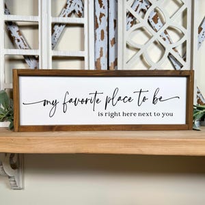May include: A rectangular wooden sign with a white background and a dark brown frame. The sign features the cursive text "my favorite place to be is right here next to you" in black lettering. The sign is displayed on a wooden shelf.