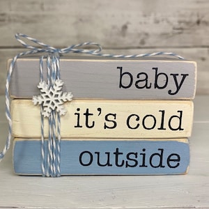 May include: A stack of three wooden blocks painted in gray, cream, and blue, tied with twine and a snowflake accent. The blocks read "baby", "it's cold", and "outside" in black lettering, evoking a winter theme.