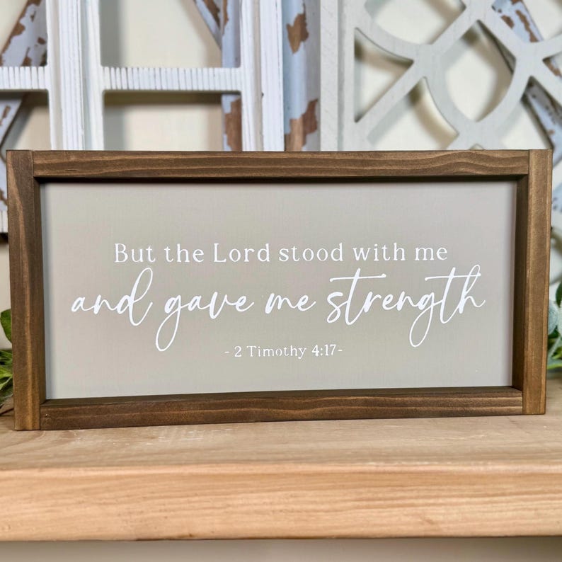 May include: A rectangular wooden framed sign with the text "But the Lord stood with me and gave me strength - 2 Timothy 4:17-" in white script. The background is a light green colour.