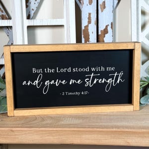 May include: A rectangular wooden framed sign with a black background. The sign features white script that reads "But the Lord stood with me and gave me strength - 2 Timothy 4:17". The frame is a light brown colour.