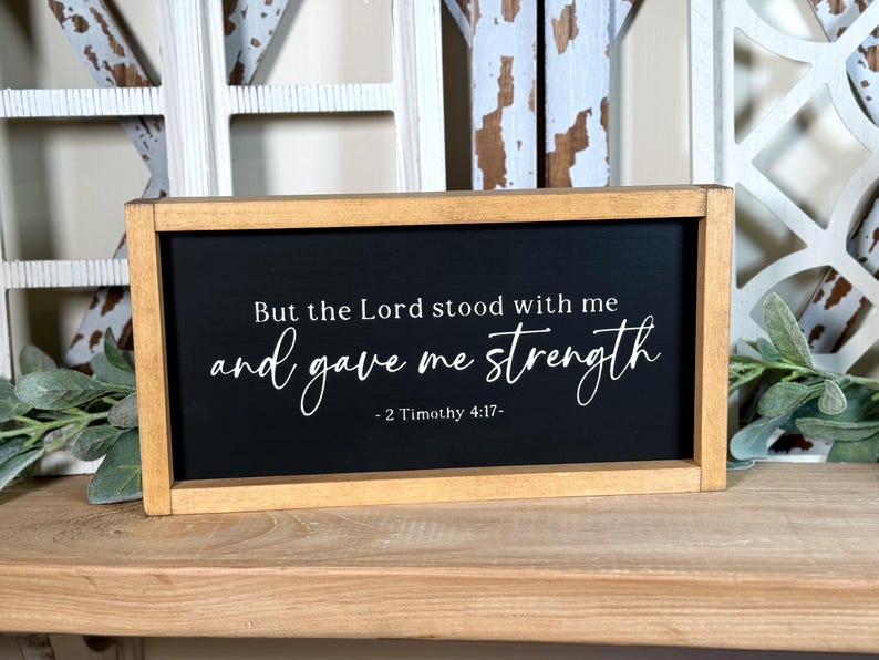 May include: A rectangular wooden framed sign with a black background. The sign features white script that reads "But the Lord stood with me and gave me strength - 2 Timothy 4:17". The frame is a light brown colour.