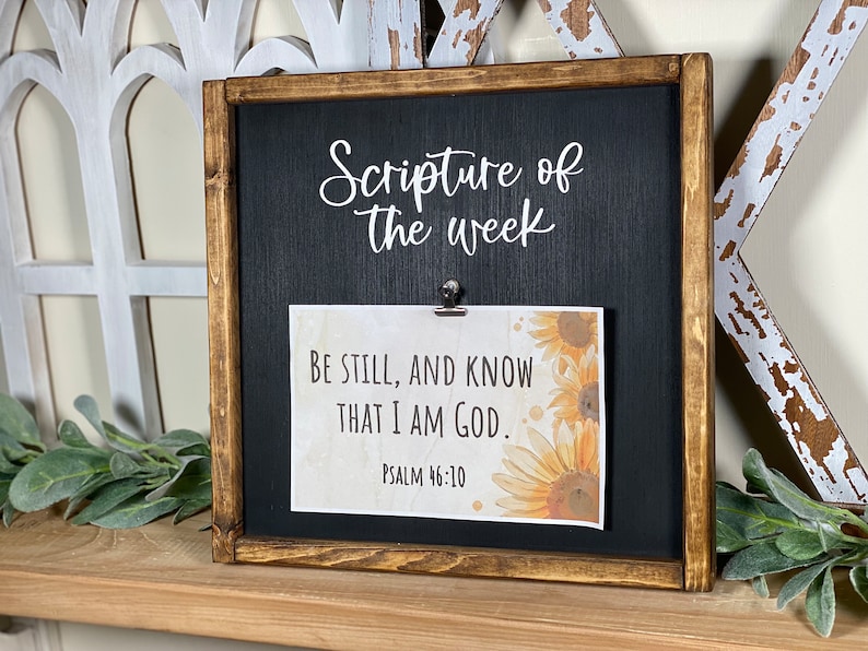 May include: A wooden frame with a black chalkboard insert and a clip to hold a card. The chalkboard says "Scripture of the week". The card has a sunflower design and the text "Be still, and know that I am God. Psalm 46:10".