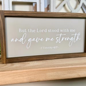 May include: A rectangular wooden sign with a dark brown frame. The sign features a light beige background with white text that reads "But the Lord stood with me and gave me strength - 2 Timothy 4:17-".
