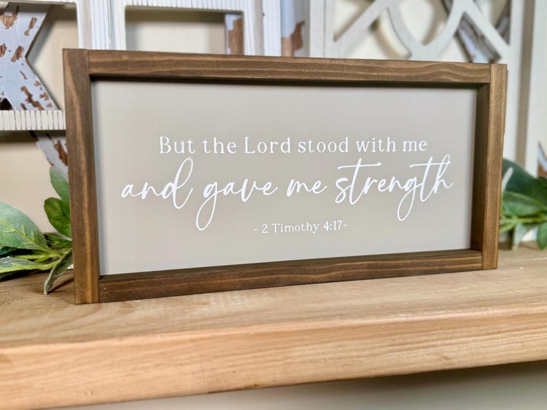 May include: A rectangular wooden sign with a dark brown frame. The sign features a light beige background with white text that reads "But the Lord stood with me and gave me strength - 2 Timothy 4:17-".
