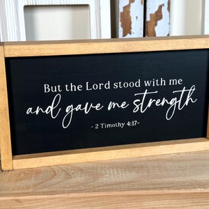 May include: Wooden framed sign with a black background. White script reads "But the Lord stood with me and gave me strength - 2 Timothy 4:17-". The frame is a light brown colour.