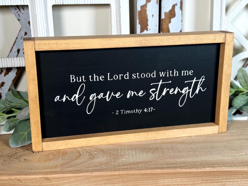 May include: Wooden framed sign with a black background. White script reads "But the Lord stood with me and gave me strength - 2 Timothy 4:17-". The frame is a light brown colour.