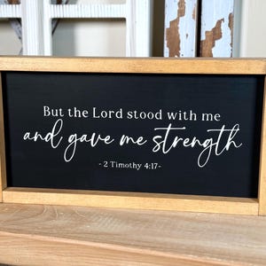May include: A framed sign with a black background and white text. The text reads "But the Lord stood with me and gave me strength - 2 Timothy 4:17-". The frame is made of light brown wood.