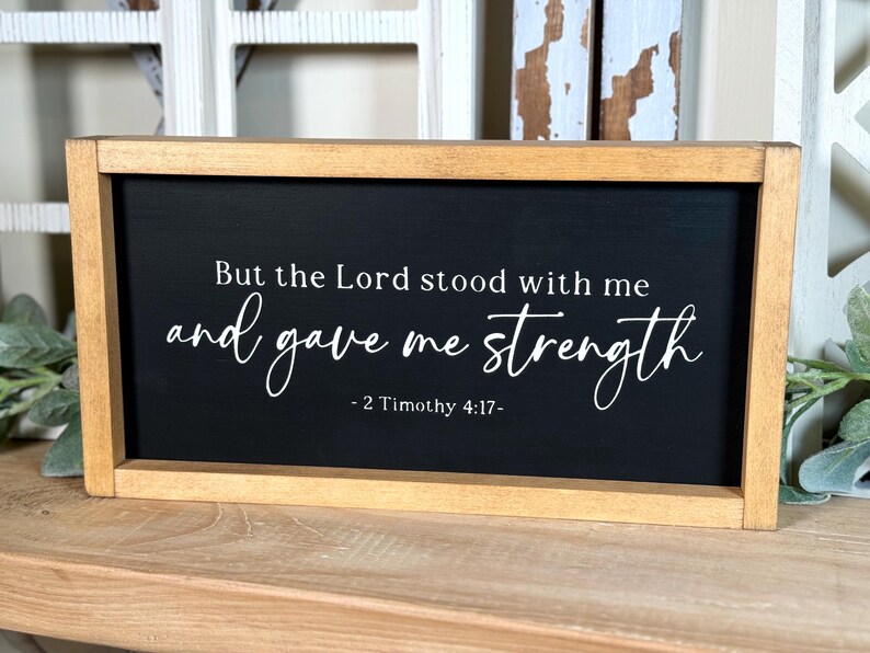 May include: A framed sign with a black background and white text. The text reads "But the Lord stood with me and gave me strength - 2 Timothy 4:17-". The frame is made of light brown wood.
