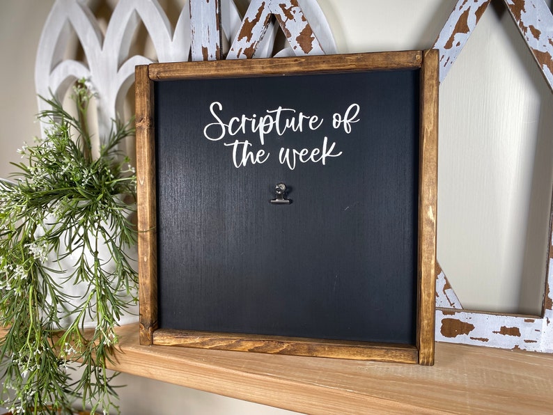 May include: A wooden framed chalkboard with the text "Scripture of the week" in white lettering. A silver binder clip is attached to the board.