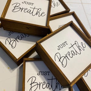 May include: A collection of square wooden signs with dark brown stained frames and white centers. Each sign features the black text "JUST Breathe" in a combination of block and script lettering. The signs are stacked in an overlapping arrangement.