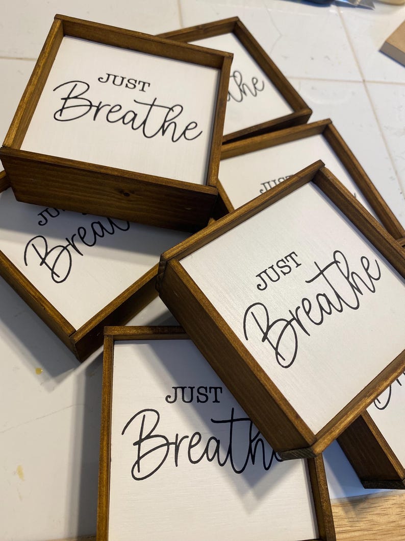 May include: A collection of square wooden signs with dark brown stained frames and white centers. Each sign features the black text "JUST Breathe" in a combination of block and script lettering. The signs are stacked in an overlapping arrangement.