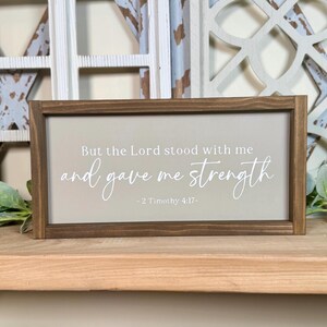 May include: A rectangular wooden sign with a light grey background and a dark brown frame. The sign features the text "But the Lord stood with me and gave me strength - 2 Timothy 4:17-" in white script. Decorative home decor.