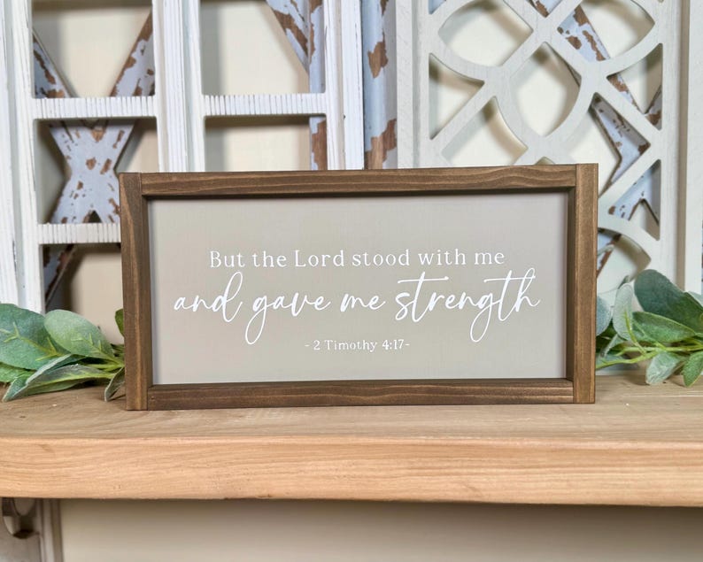 May include: A rectangular wooden sign with a light grey background and a dark brown frame. The sign features the text "But the Lord stood with me and gave me strength - 2 Timothy 4:17-" in white script. Decorative home decor.