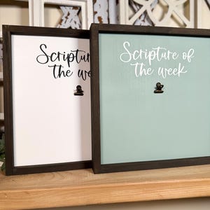 May include: Two wooden framed signs with black frames and a clip on each. The sign on the left is white with black text that says "Scripture of the week". The sign on the right is light blue with white text that says "Scripture of the week".