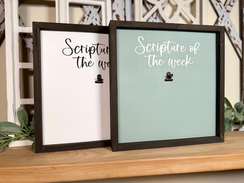 May include: Two wooden framed signs with black frames and a clip on each. The sign on the left is white with black text that says "Scripture of the week". The sign on the right is light blue with white text that says "Scripture of the week".