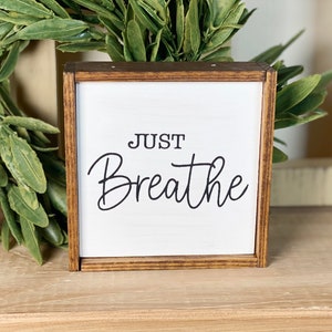 May include: A square wooden sign with a dark brown stained frame and a white center panel. The text "JUST Breathe" is printed in black, with "JUST" in a simple sans-serif font and "Breathe" in a flowing, cursive script typeface.