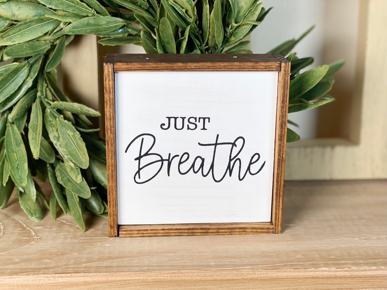 May include: A square wooden sign with a dark brown stained frame and a white center panel. The text "JUST Breathe" is printed in black, with "JUST" in a simple sans-serif font and "Breathe" in a flowing, cursive script typeface.