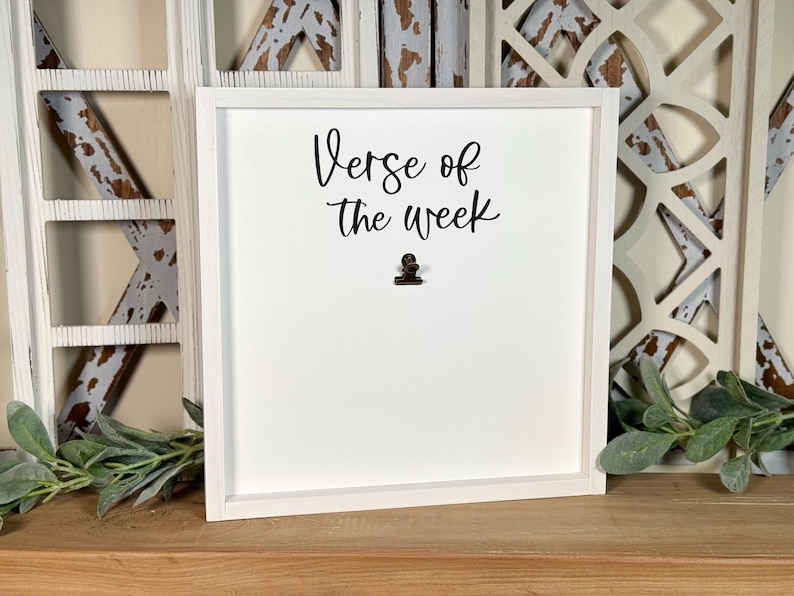 May include: White framed sign with the text "Verse of the week" in black cursive lettering. A small metal clip is attached to the sign.