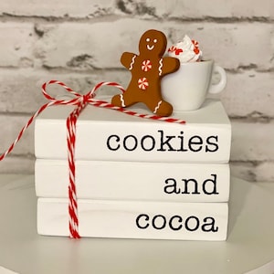 May include: A stack of three white books tied with red and white twine. The books read "cookies and cocoa" in black text. A gingerbread man and a mug of whipped cream and candy are on top. The background is a white brick wall.