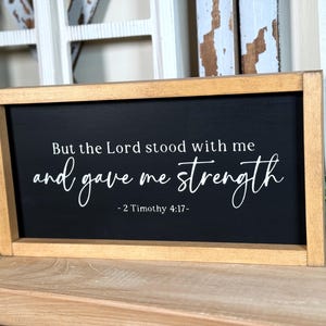 May include: Rectangular wooden sign with a black surface and white lettering. The text says "But the Lord stood with me and gave me strength - 2 Timothy 4:17-". The frame is light brown. Suitable for home decoration.