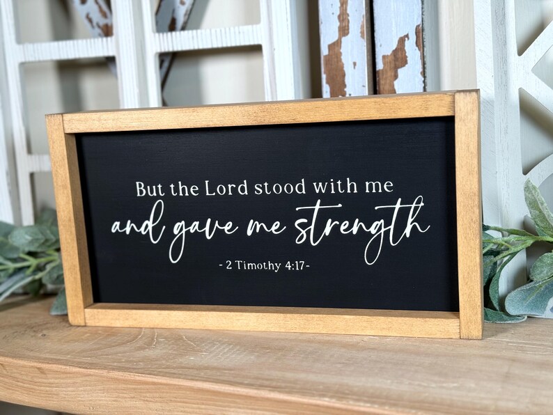 May include: Rectangular wooden sign with a black surface and white lettering. The text says "But the Lord stood with me and gave me strength - 2 Timothy 4:17-". The frame is light brown. Suitable for home decoration.