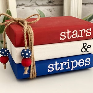 May include: A stack of three wooden books painted red, white, and blue, tied with twine and adorned with red and blue polka dot beads. The top book reads "stars", the middle book has an ampersand, and the bottom book reads "stripes".