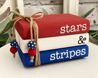 Peut inclure: Une pile de trois livres en bois peints en rouge, blanc et bleu, attachés avec de la ficelle et ornés de perles à pois rouges et bleues. Les livres affichent les mots "stars", un esperluette, et "stripes".