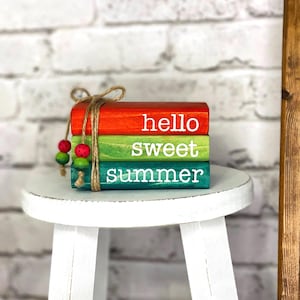 May include: A stack of three wooden blocks painted in bright colors: red, green, and teal. The blocks are tied together with twine and adorned with decorative beads. Each block has a white word: "hello", "sweet", and "summer". The blocks are on a white stool.