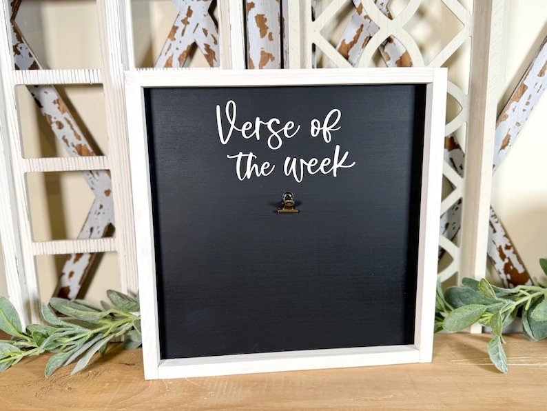 May include: White framed chalkboard with the text "Verse of the week" in white cursive lettering. A small gold clip is attached to the board.