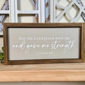 May include: A rectangular wooden sign with a dark brown frame. The sign has a light beige background with the text "But the Lord stood with me and gave me strength - 2 Timothy 4:17-" in white script.