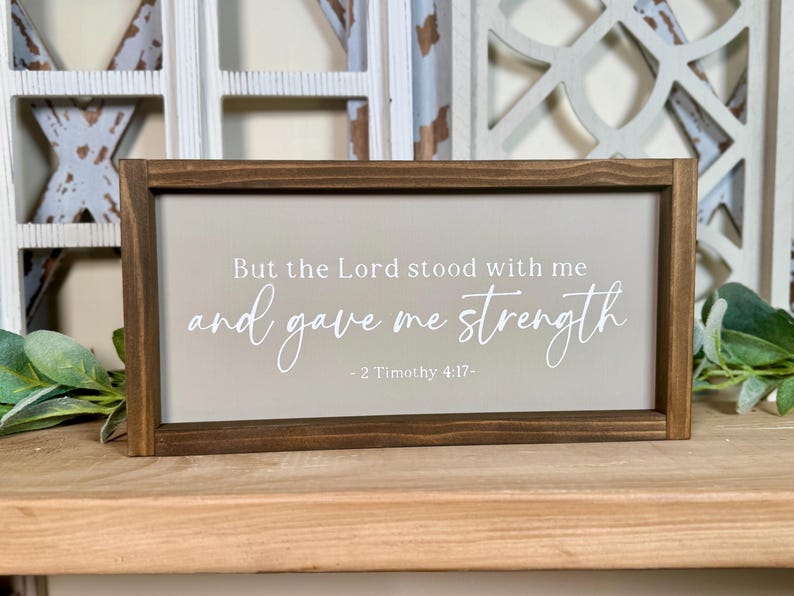May include: A rectangular wooden sign with a dark brown frame. The sign has a light beige background with the text "But the Lord stood with me and gave me strength - 2 Timothy 4:17-" in white script.