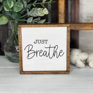 May include: A square wooden sign with a white face and a dark brown stained frame. The text "JUST Breathe" is printed in black lettering on the front. The sign features a clean, simple design with a smooth finish and visible wood grain texture.