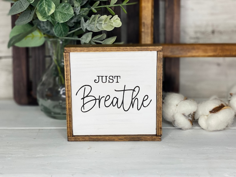 May include: A square wooden sign with a white face and a dark brown stained frame. The text "JUST Breathe" is printed in black lettering on the front. The sign features a clean, simple design with a smooth finish and visible wood grain texture.