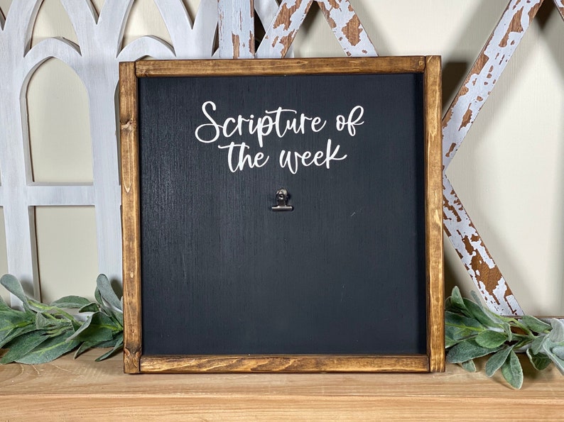 May include: A dark brown wooden framed chalkboard with the text "Scripture of the week" in white lettering. A silver clip is attached to the board.