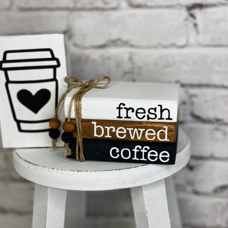 Może przedstawiać: Trzy ułożone na sobie drewniane klocki z napisem "fresh brewed coffee" czarnymi literami. Klocki są pomalowane na biało, brązowo i czarno. Klocki są związane sznurkiem i mają przymocowane drewniane koraliki.