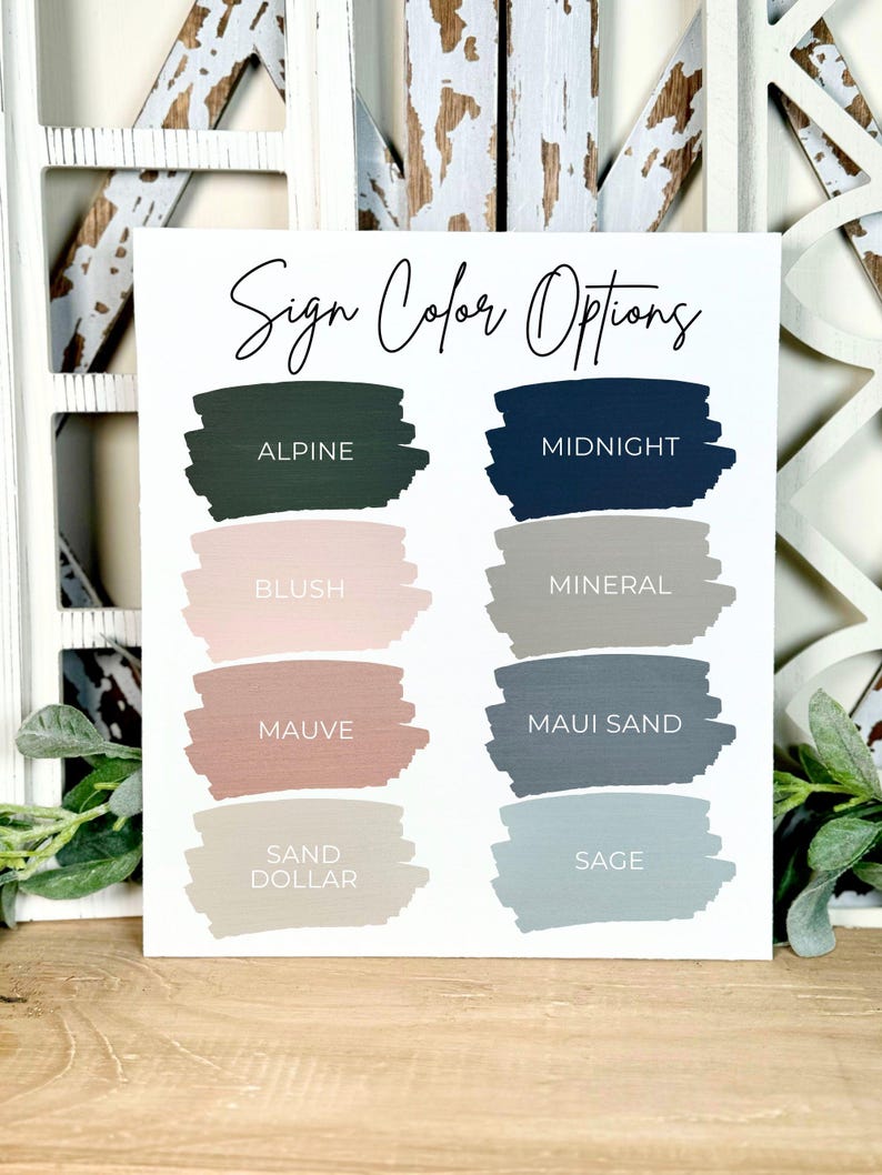 May include: A white sign with the text "Sign Color Options" in script. Below, nine paint swatches display colour choices including Alpine, Midnight, Blush, Mineral, Mauve, Maui Sand, Sand Dollar, and Sage. The sign is set against a rustic, white-washed wooden background.
