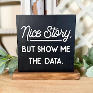 May include: Black chalkboard sign with a white wooden base. The sign reads "Nice story, but show me the data."
