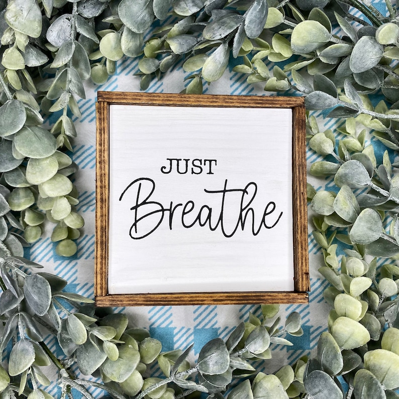 May include: A square wooden sign with a white background and a dark brown stained frame. The text "JUST Breathe" is printed in black lettering on the center of the sign. The word "JUST" is in a simple sans-serif font, while "Breathe" is in script.