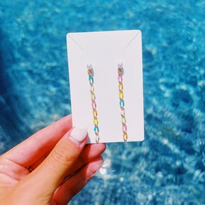 May include: A pair of long drop earrings featuring a small white pearl stud at the top, connected to a dangling chain of multicolored oval links in shades of pink, yellow, blue, and white, displayed on a plain white rectangular card.