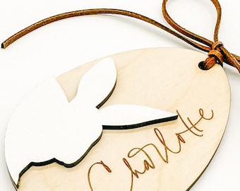 May include: A wooden Easter egg ornament with a white bunny cutout and the name "Charlotte" engraved on it. The ornament is attached to a brown leather cord.