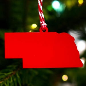 May include: Red Christmas ornament shaped like the state of Nebraska hanging from a red and white striped string on a green Christmas tree.