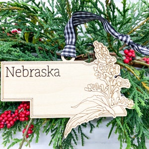 May include: Wooden ornament shaped like the state of Nebraska with a floral design and the word "Nebraska" inscribed on the top. The ornament is hanging from a black and white ribbon.