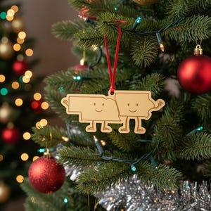 May include: A wooden Christmas ornament shaped like two states, each with a smiling face and arms raised. The ornament hangs from a red ribbon on a decorated Christmas tree with red and gold ornaments and lights.