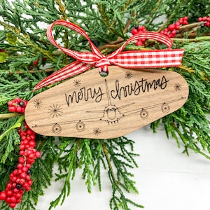 May include: Wooden Christmas ornament shaped like an airplane with the text "merry christmas" on it. The ornament is decorated with a red and white gingham ribbon.