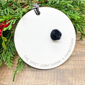 May include: White round ornament with black text that reads "The Rivet Joint During A Snowstorm". A black pom-pom is attached to the ornament.
