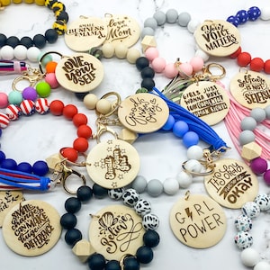 May include: A collection of wooden keychains with various inspirational messages, including "Love Yourself", "Small Town Girl", "Good Vibes Only", "Girl Power", and "It's Okay to Not Be Okay". The keychains are made of natural wood and feature a variety of colors and patterns.